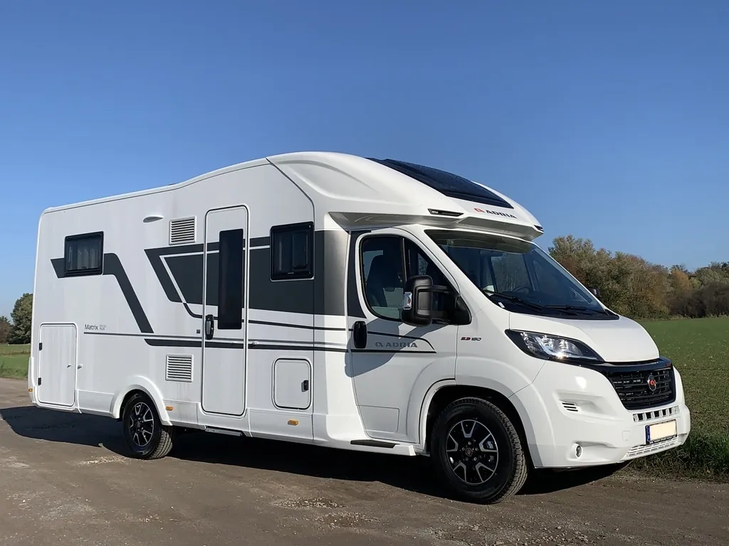 Autocaravana moderna blanca estacionada en un camino, con un claro cielo azul de fondo y áreas verdes a los lados